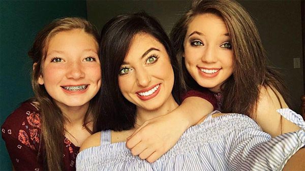 The image shows three individuals posing for a photograph with smiles on their faces two girls are hugging each other while a woman stands behind them, all appear to be indoors with a blue wall visible in the background.